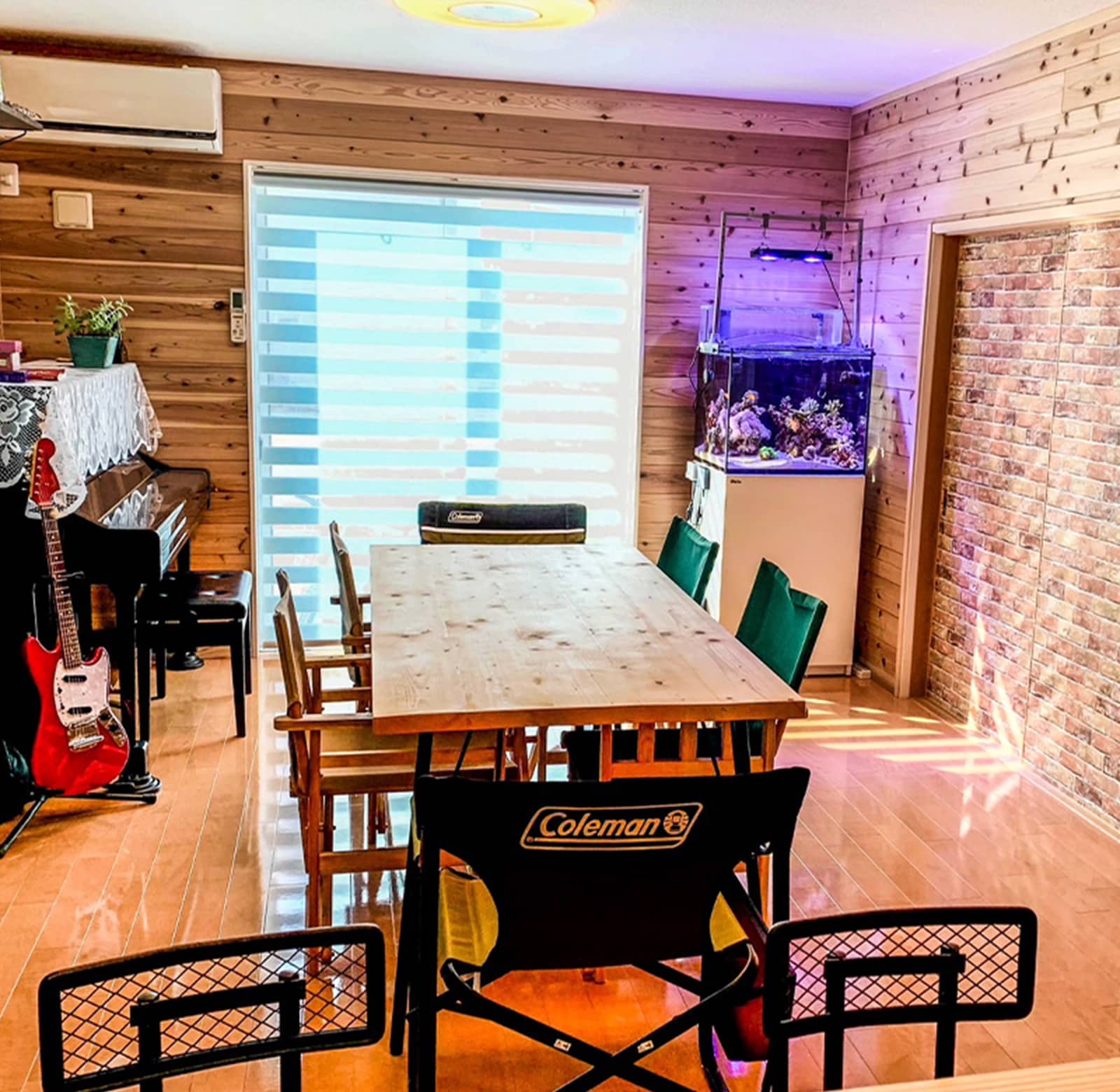 木製のテーブルと椅子が置かれた室内。部屋の奥にはピアノやギター、水槽が設置されている。
